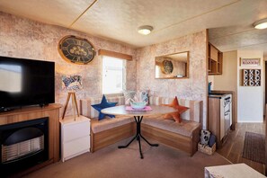A compact living space with a television, a clock, a dining table, and a kitchenette.