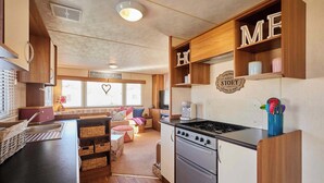 A compact kitchen with a stove, sink, and wooden cabinets, a cozy seating area with cushions, and a window with blinds. - Host & Stay | Cosy Cove (Cresswell)