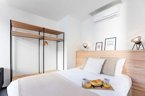 A modern hotel room with a wooden headboard, a bed with white linens, a breakfast tray with croissants and orange juice, and a wall-mounted air conditioner. - Regras Apartments & Garden A11 by Benefit Collection (Porto)