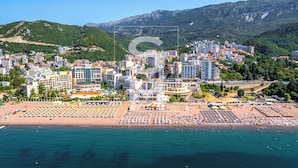 A coastal cityscape with a beach area featuring numerous sunbeds and umbrellas, surrounded by residential buildings and mountains in the background. - Status apartments 173 (Becici)