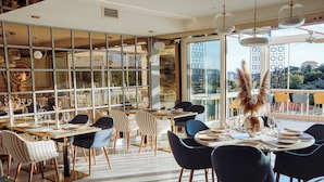 A modern dining area with tables set for a meal, large windows offering a view of the city, and a decorative plant centerpiece. - Status apartments 173 (Becici)
