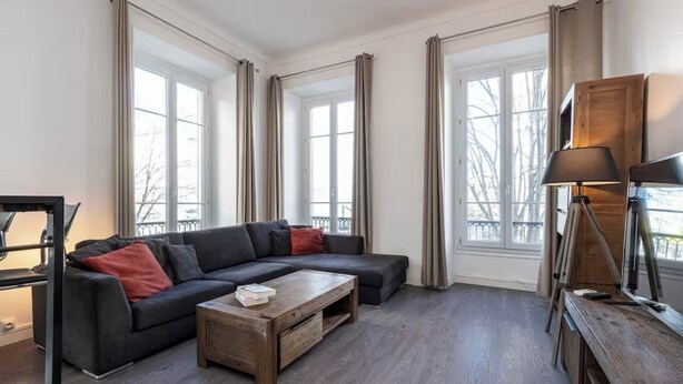 Un salon moderne avec un canapé d’angle, une table basse en bois et un bureau avec une lampe. De grandes fenêtres laissent entrer la lumière naturelle et offrent une vue sur l’extérieur.