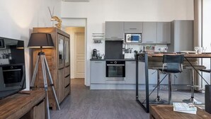 Une cuisine moderne avec des meubles gris, un four encastré et un micro-ondes. On y trouve également un coin bar avec des tabourets et une télévision posée sur une table en bois.