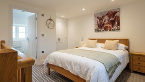 Une chambre à coucher avec un lit en bois, du linge de lit blanc, une table de chevet, une chaise en bois, une porte donnant sur un balcon et un tableau représentant des bovins des Highlands accroché au mur.