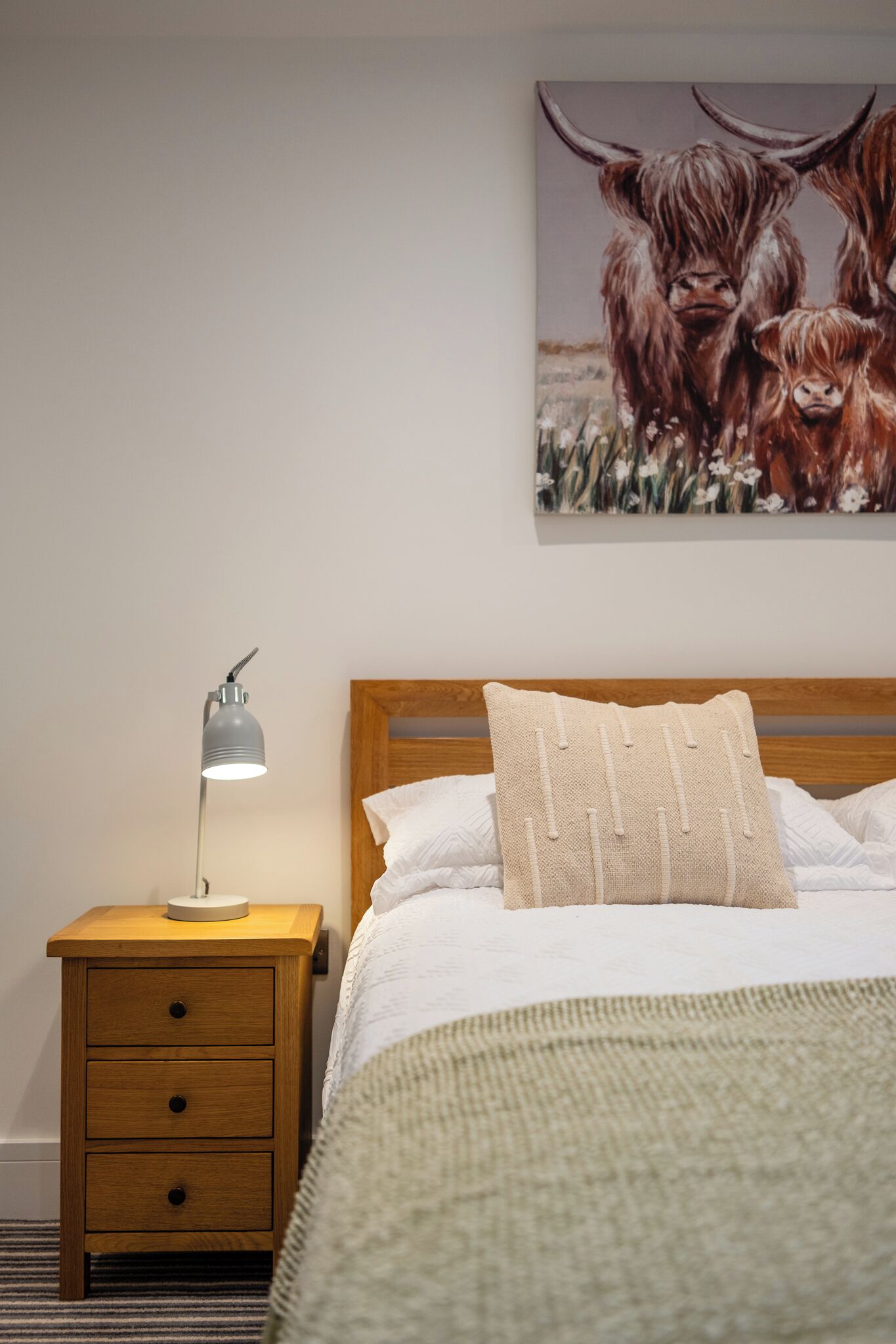 Un dormitorio con una mesita de noche de madera, una lámpara, una cama con ropa de cama blanca y un cuadro de yakas en la pared.