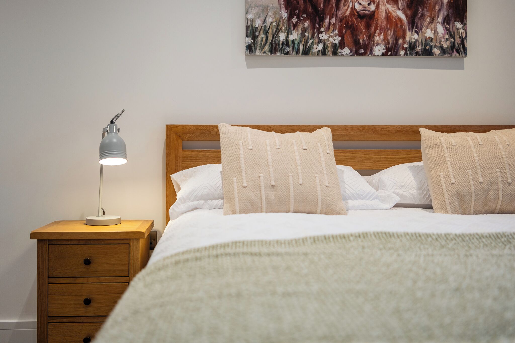 A neatly made bed with white linens and beige pillows, a bedside lamp, and a wooden nightstand with drawers.