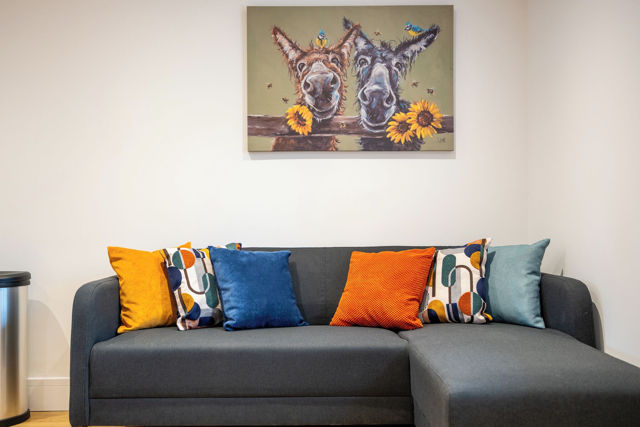 Un sofá seccional gris con cojines de colores y estampados, un cuadro de burros y girasoles en la pared, y un cubo de basura a la izquierda.