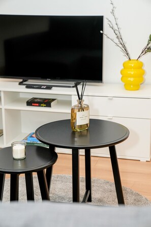 Una sala moderna con un televisor de pantalla plana, una pequeña mesa redonda con una vela y un jarrón decorativo sobre un mueble blanco.