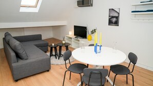 A modern living room with a grey sectional sofa, a round white table with chairs, a flat-screen TV, a small side table with candles, and a picture on the wall. - Modern 1BR 1LR Apartment Walk To Rabot (Ghent)