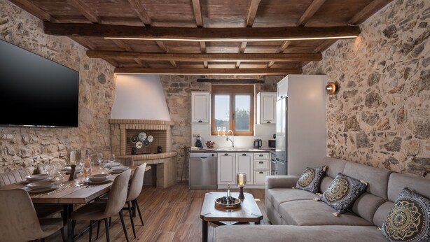 A cozy living room with a fireplace, a dining area, and a kitchen with stone walls and wooden beams. - Stella s Traditional House Near the City (Chania)