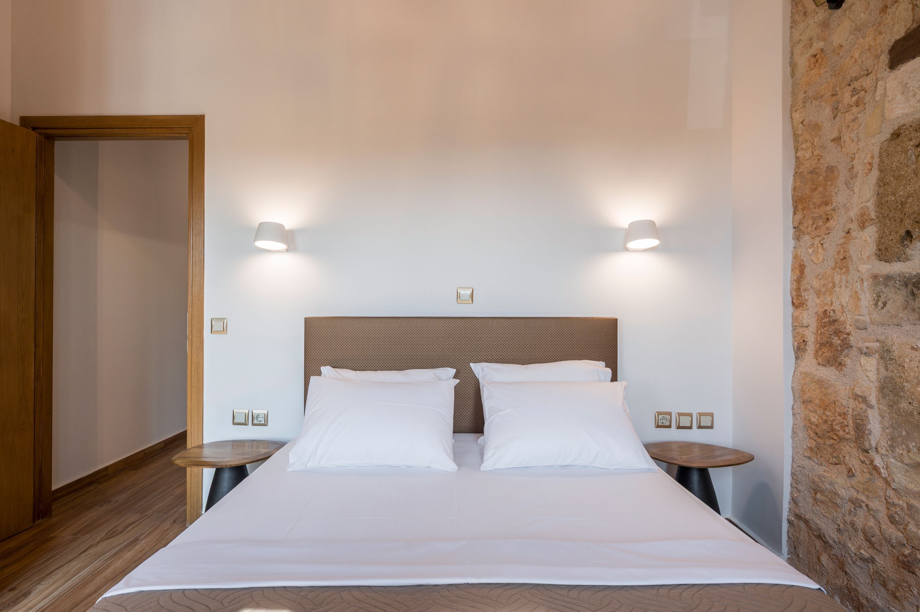 A neatly made bed with white linens, two bedside tables with lamps, and a stone wall to one side.