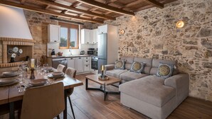 A cozy living area with a stone wall, wooden ceiling beams, a sofa set, and a dining table set for six. - Stella s Traditional House Near the City (Chania)