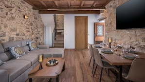 A cozy living area with a stone wall, wooden ceiling beams, a sofa with patterned cushions, a dining table set for six, and a staircase leading to another room. - Stella s Traditional House Near the City (Chania)
