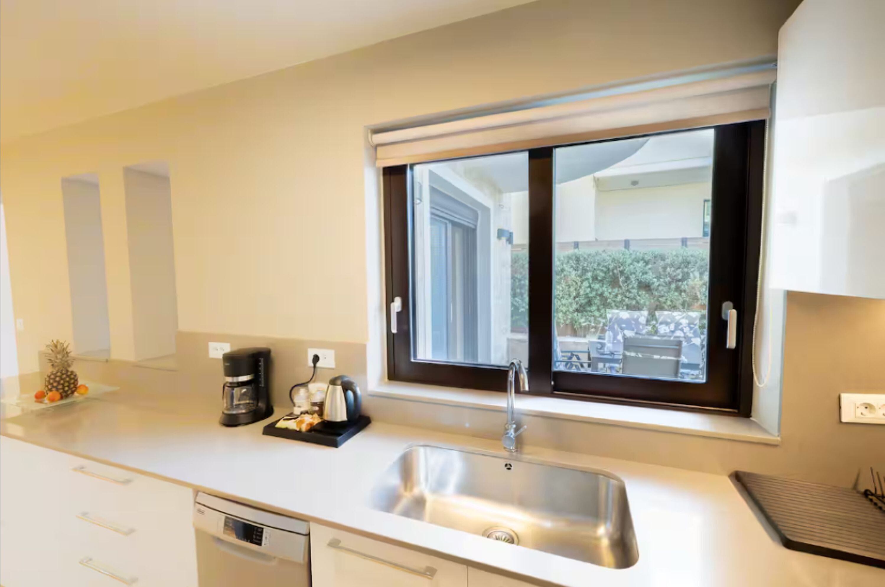 A modern kitchen with a stainless steel sink, a window with blinds, and a countertop with a coffee maker and fruit.