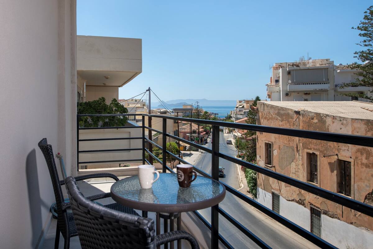 A balcony with wicker chairs and a small table, overlooking a street with buildings and a clear view of the sea.