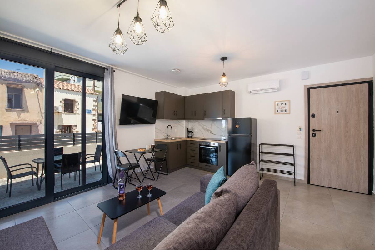 A modern apartment with a kitchen, dining area, and living room. It features a large sliding glass door to the outside, a flat-screen TV mounted on the wall, and a cozy seating area with a sofa and a small table.