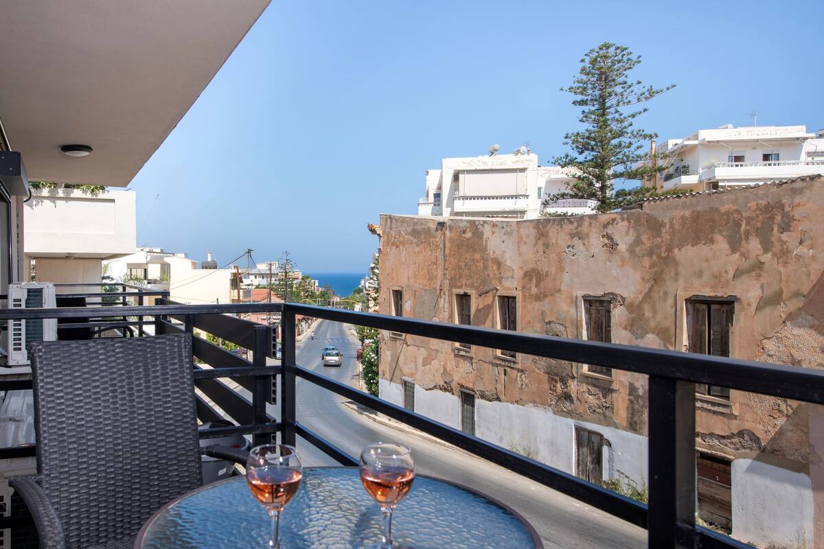 A balcony with two wine glasses on a round table, overlooking a street with buildings and a clear sky.