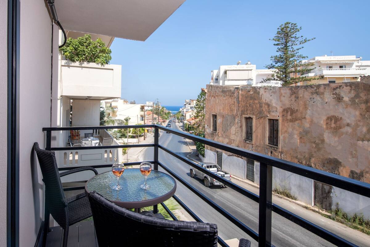 A balcony with a table and chairs, overlooking a street with buildings and a clear sky.