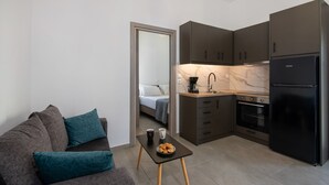 A compact living space with a kitchenette, a sofa with teal cushions, a small table with a coffee cup and croissant, and a bed in the adjacent room. - Halepa Apartment's in Chania (Chania)
