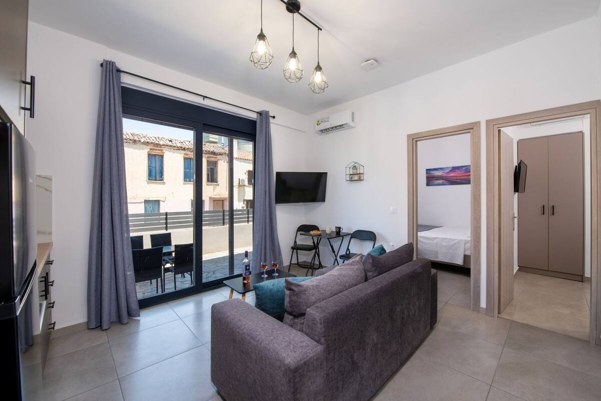 A modern living room with a sofa, a TV, a dining table, and a balcony with a view of buildings.