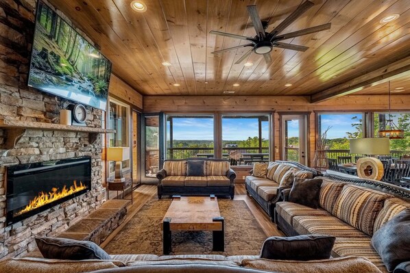 A spacious living room with a fireplace, large windows, a ceiling fan, and a television mounted on a stone wall.