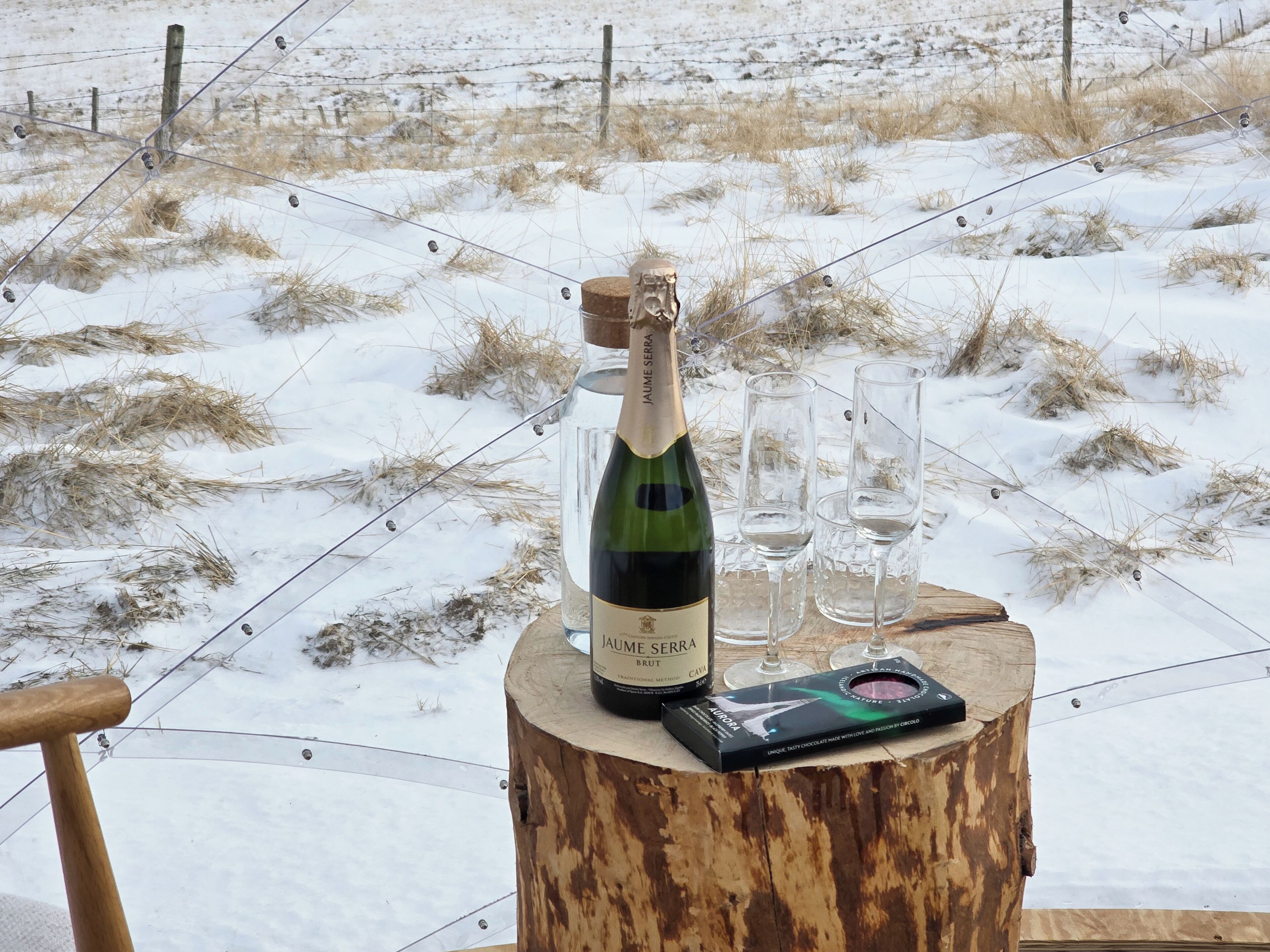 Una botella de vino Jaume Serra Brut, dos copas y una caja de bombones sobre un tocón de madera en un entorno nevado al aire libre.