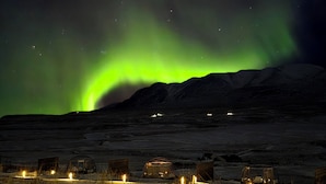 A remote, snow-covered landscape with several small, rustic cabins under a vibrant green aurora display.