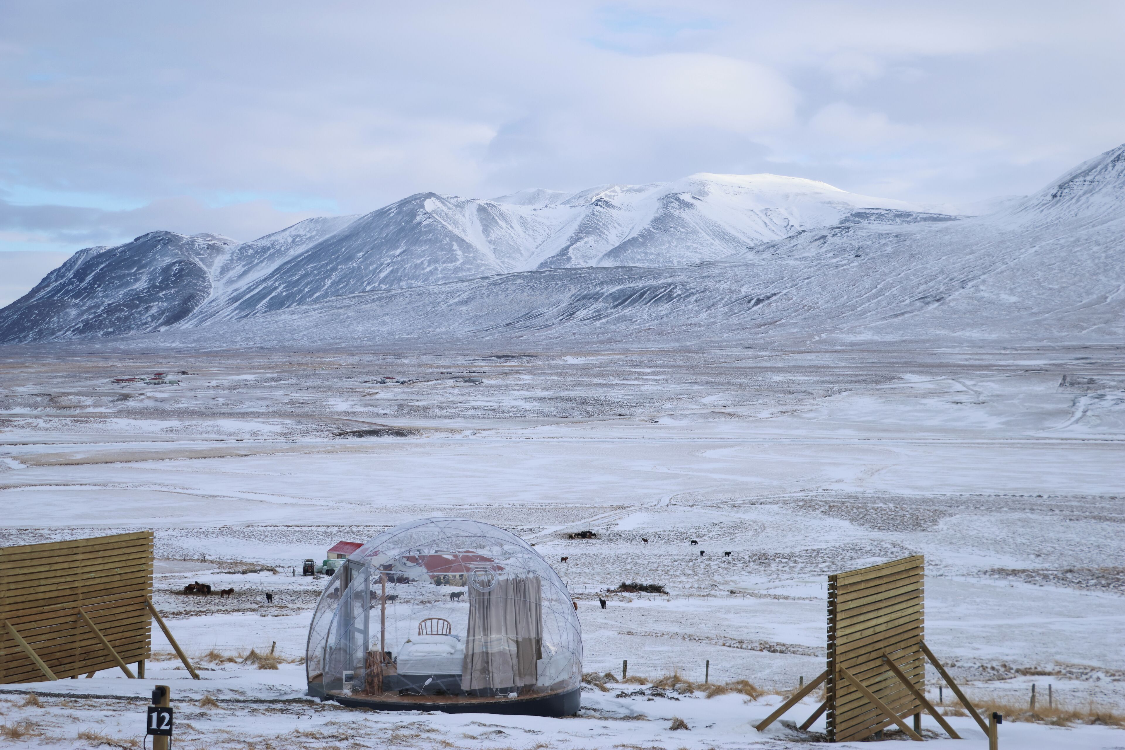 A transparent dome structure with a bed and a small table, surrounded by snow-covered ground and mountains in the background.