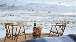 Un coin salon accueillant avec deux chaises en bois et une petite table en bois, agrémenté d’une bouteille et d’un verre, donnant sur un paysage enneigé avec des montagnes lointaines.