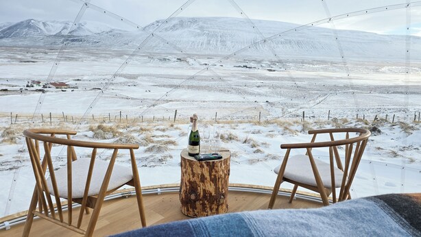 A cozy seating area with two wooden chairs and a small wooden table, featuring a bottle and glass, overlooking a snowy landscape with distant mountains.