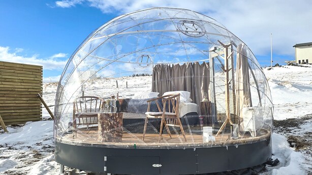 Une structure en forme de dôme transparent, avec une terrasse en bois, des chaises et un lit à l’intérieur.