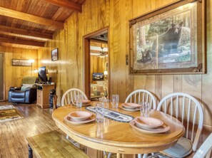 A wooden dining area with a round table set for four, surrounded by white chairs. A framed wildlife picture hangs on the wall, and a television is visible in the background. - Bear Cub Retreat - A charming cabin less than a mile to Gatlinburg! (Gatlinburg)