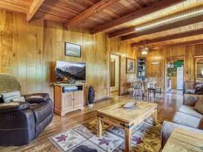 A cozy living room with a wooden ceiling, a flat-screen TV, leather sofa, and a dining area with chairs and a table. - Bear Cub Retreat - A charming cabin less than a mile to Gatlinburg! (Gatlinburg)