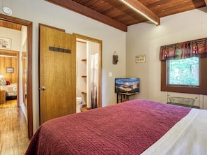 A bedroom with a wooden door, a bed with a red quilt, a television on the wall, and a window with a view of trees. - Bear Cub Retreat - A charming cabin less than a mile to Gatlinburg! (Gatlinburg)