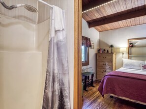 Ein Badezimmer mit Dusche, Holzboden, einem Bett mit violetter Bettdecke, einem Schminktisch und einem Fenster mit Blick auf Bäume.