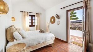 A bedroom with a bed, a wicker chair, a window with curtains, and a view of the outdoors. - Casa del Mar. Spectacular villa with sea views and jacuzzi in Charco del Palo (Chraco del Palo - Haría)