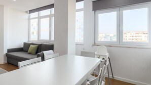 A modern, well-lit room with a white table, grey sofa, and a view of the city through large windows. - Apartment 'M J House' with Sea View, Wi-Fi and Air Conditioning (Setúbal)