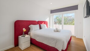 A bedroom with a red headboard, white bedding, a bedside lamp, and a TV mounted on the wall. - Apartment 'M J House' with Sea View, Wi-Fi and Air Conditioning (Setúbal)