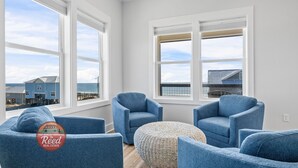 Een kamer met drie blauwe fauteuils, een rieten poef en grote ramen met uitzicht op de oceaan.
