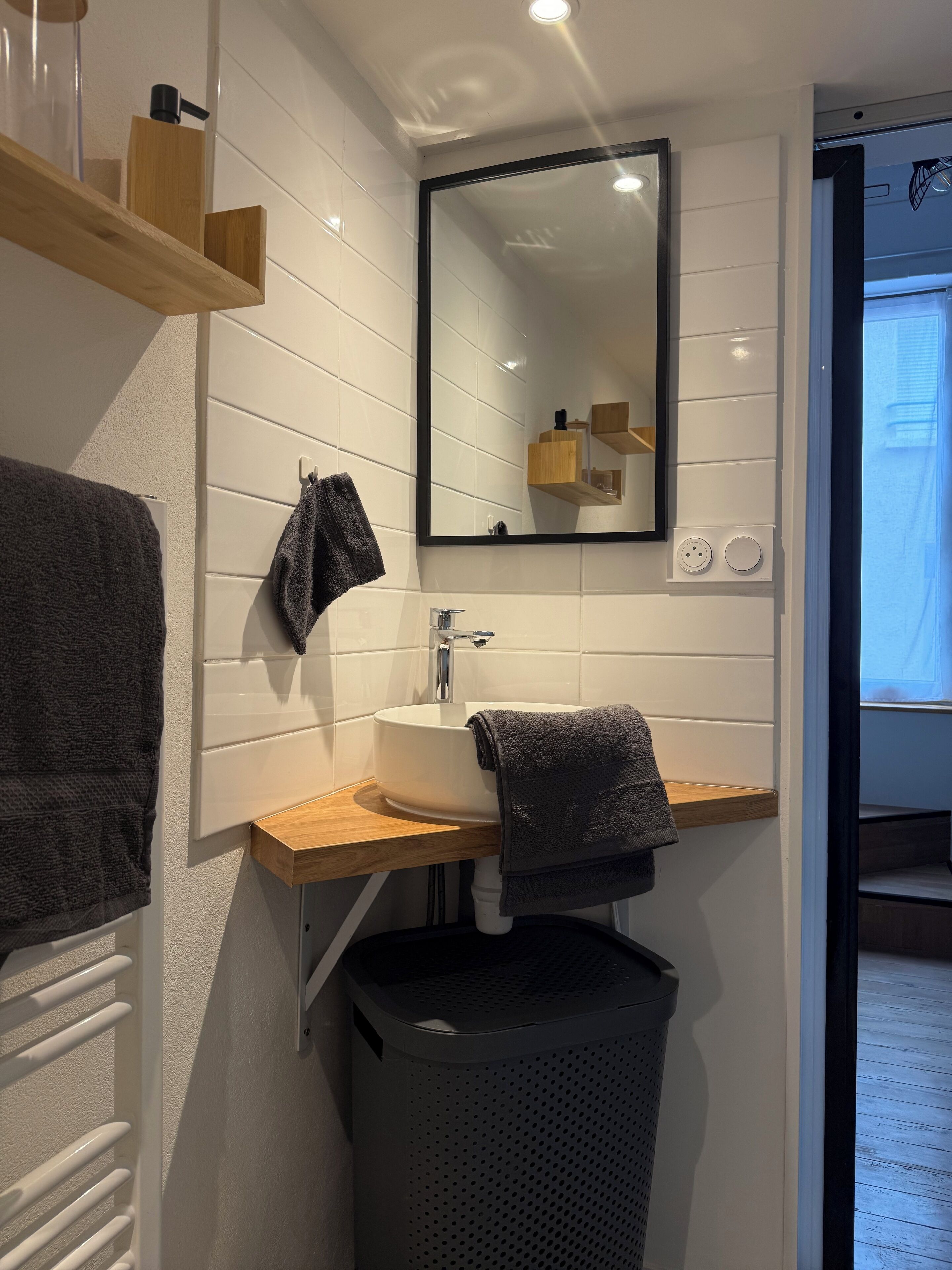 Une salle de bain avec un lavabo blanc, un miroir et des étagères en bois. On y trouve un porte-serviettes avec une serviette et une poubelle.