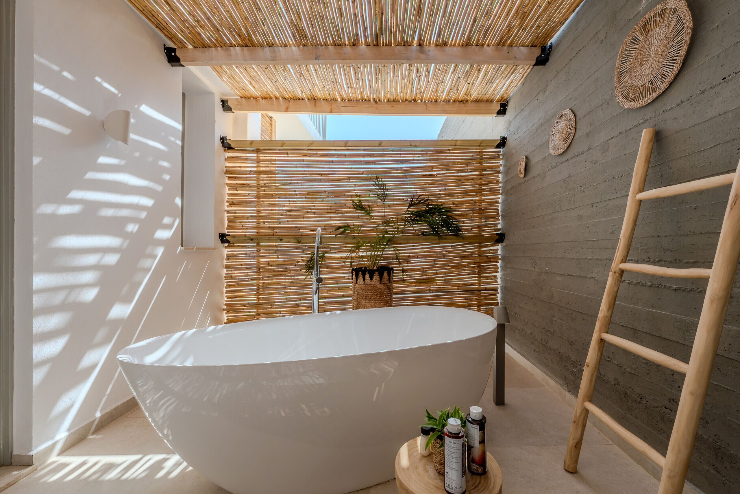 Un baño moderno con una bañera independiente grande, techo de bambú y pared de concreto con cestas tejidas.