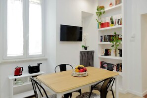Zona pranzo con tavolo in legno, sedie, una ciotola di frutta, televisione montata a parete e una libreria con libri e piante.