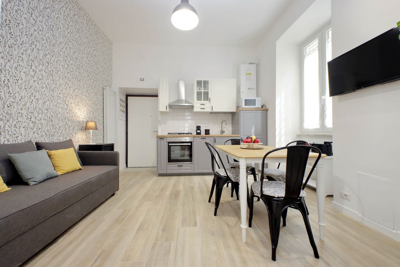 A modern kitchen with white cabinets, a microwave, and an oven. There is a dining table with chairs and a sofa with cushions.