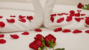 A bed with white linens, two towels folded into heart shapes, and scattered red rose petals.