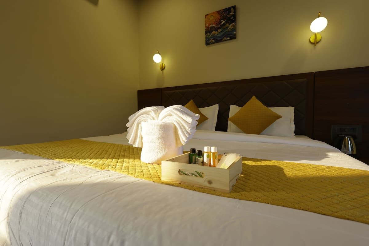 Una cama bien hecha con un edredón amarillo, una mesita de noche de madera con artículos de aseo, y dos lámparas de pared.