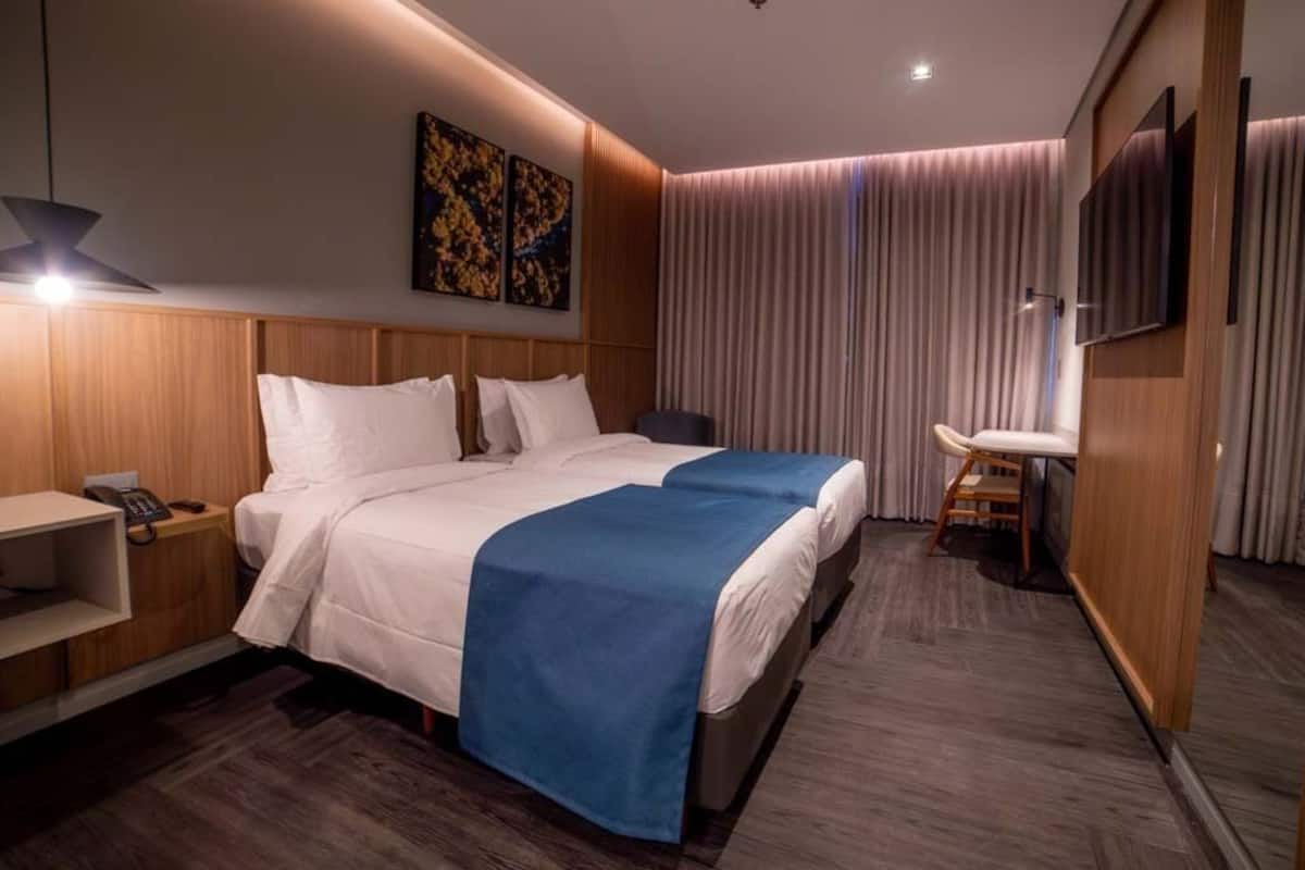 A hotel room with a large bed, a bedside table with a phone, a small desk with a chair, a TV mounted on the wall, and a window with curtains.