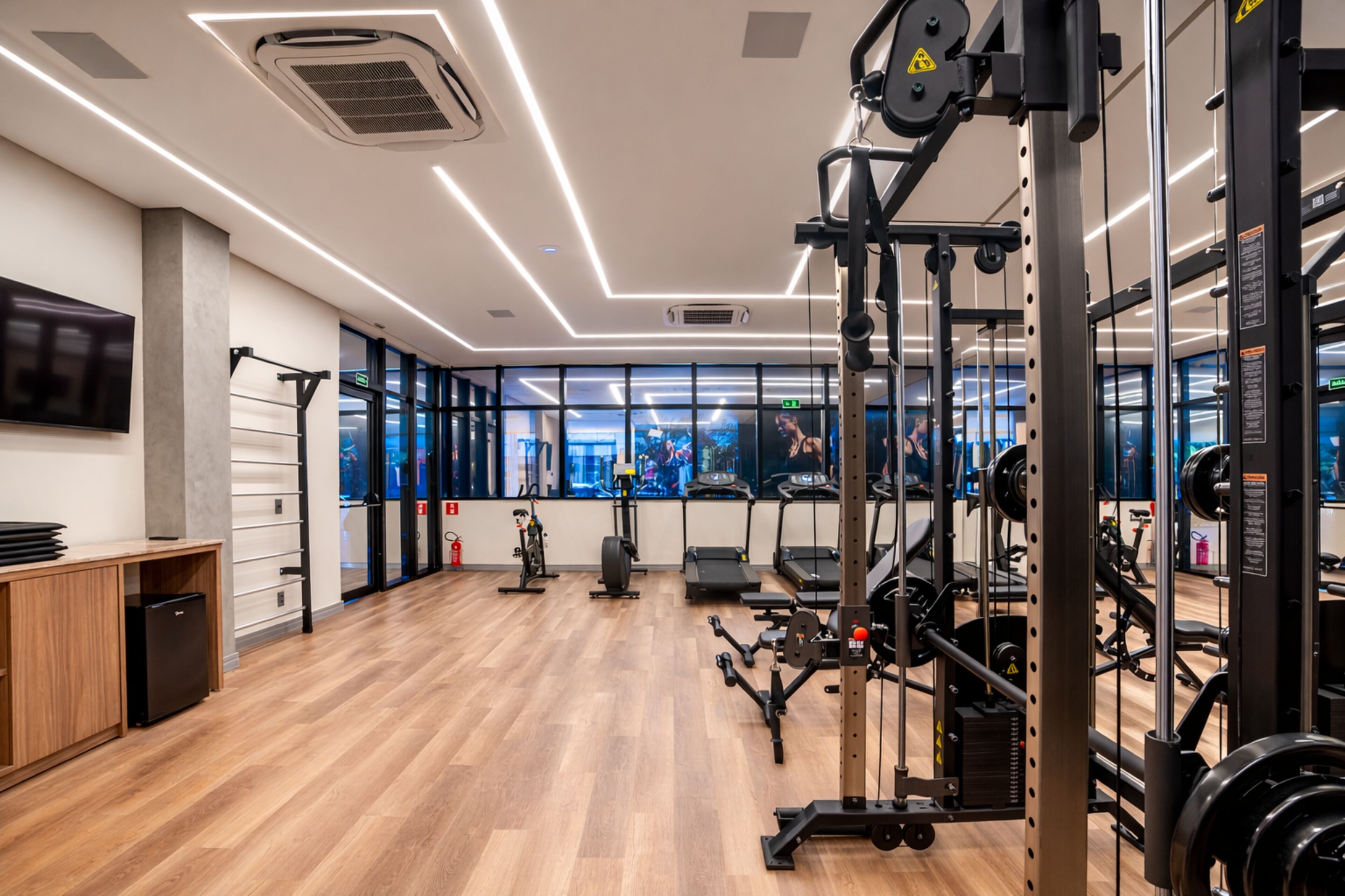 A modern gym with exercise equipment, a wooden floor, and large windows.