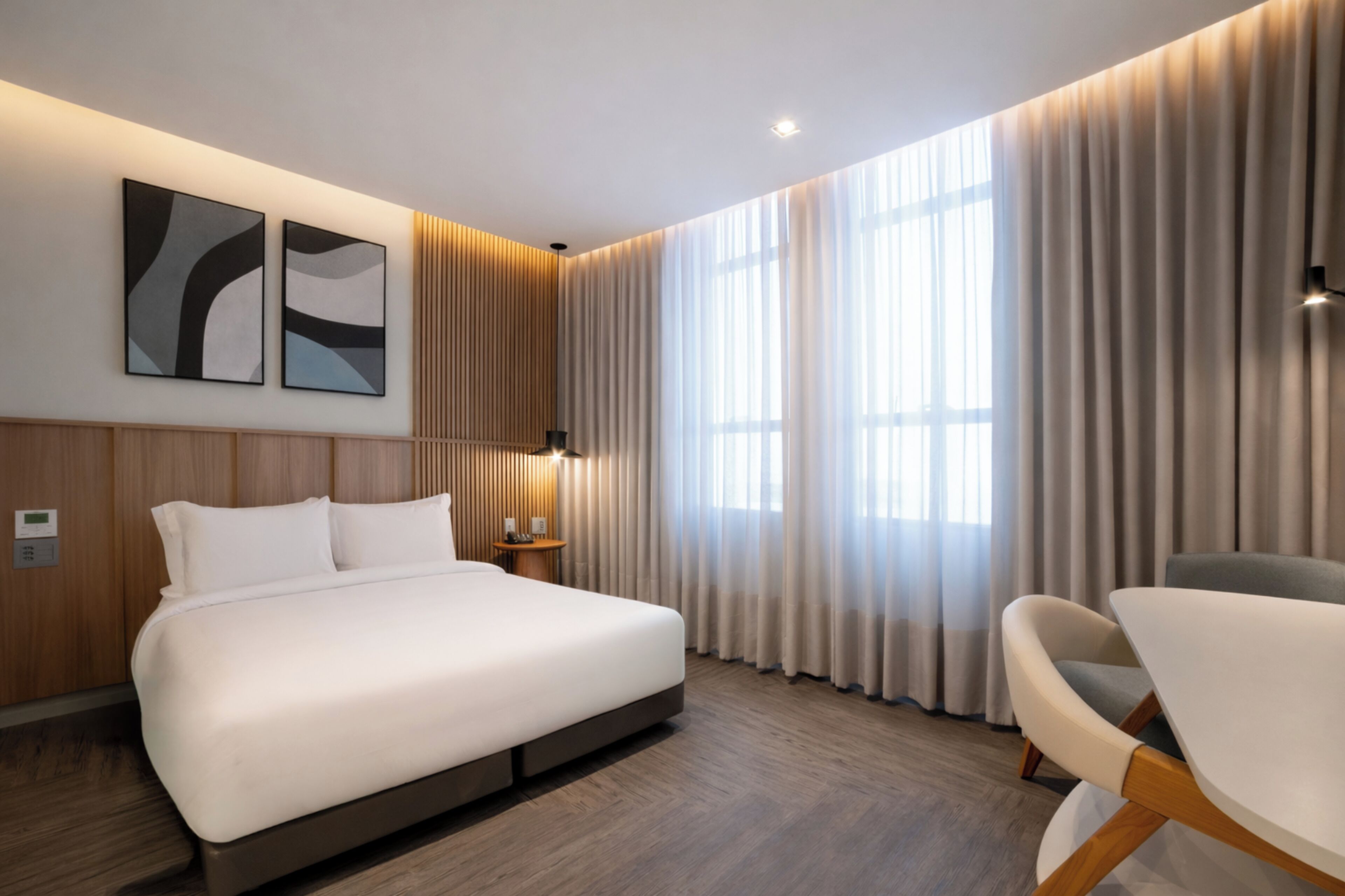 A modern hotel room with a large bed, a bedside table, a chair, and a large window with curtains.