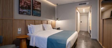 A modern hotel room with a bed, bedside tables, a small seating area, and a hallway leading to a bathroom.