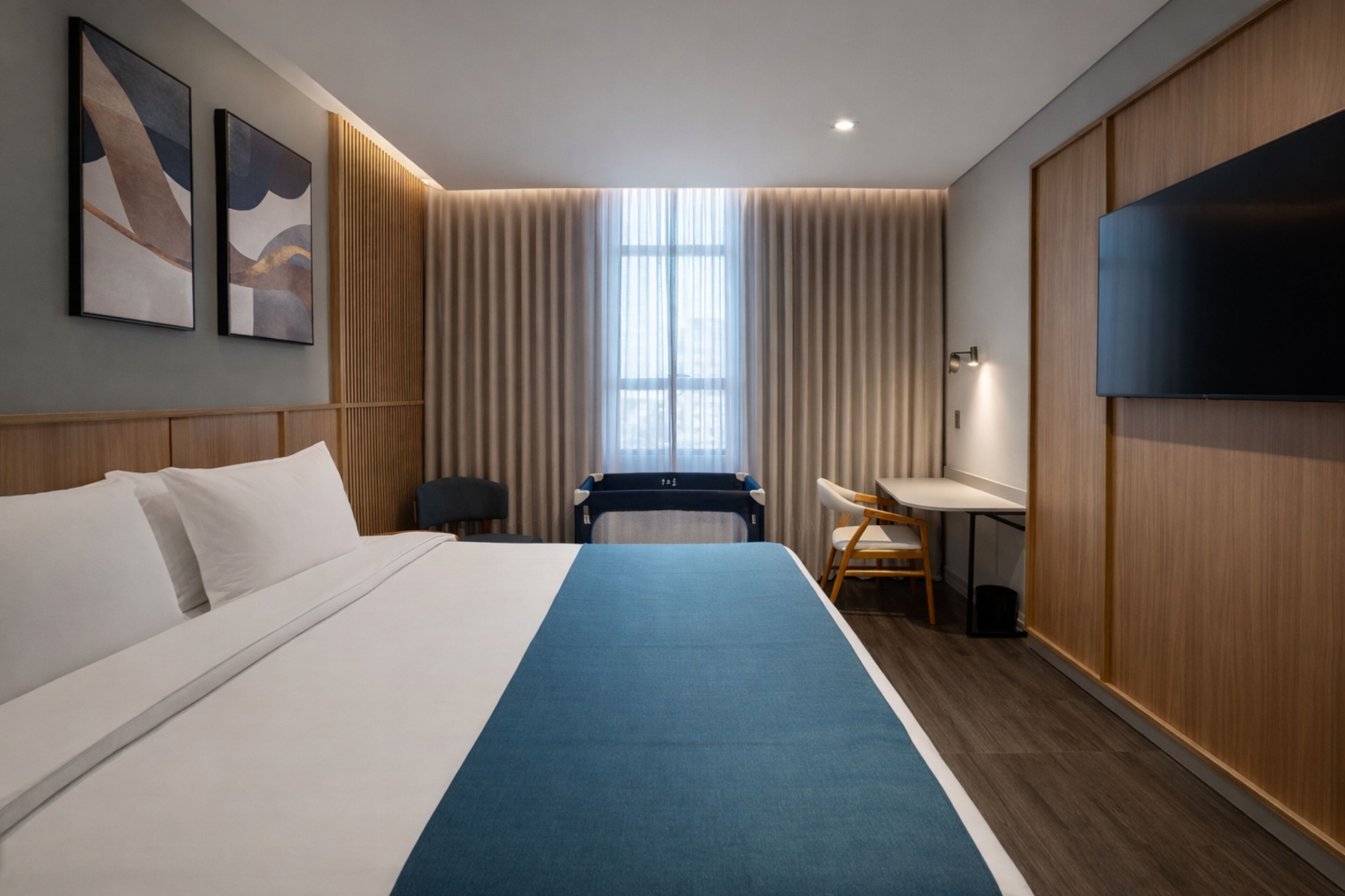 Quarto de hotel com uma cama grande, uma escrivaninha, uma cadeira, televisão e janela com cortinas.